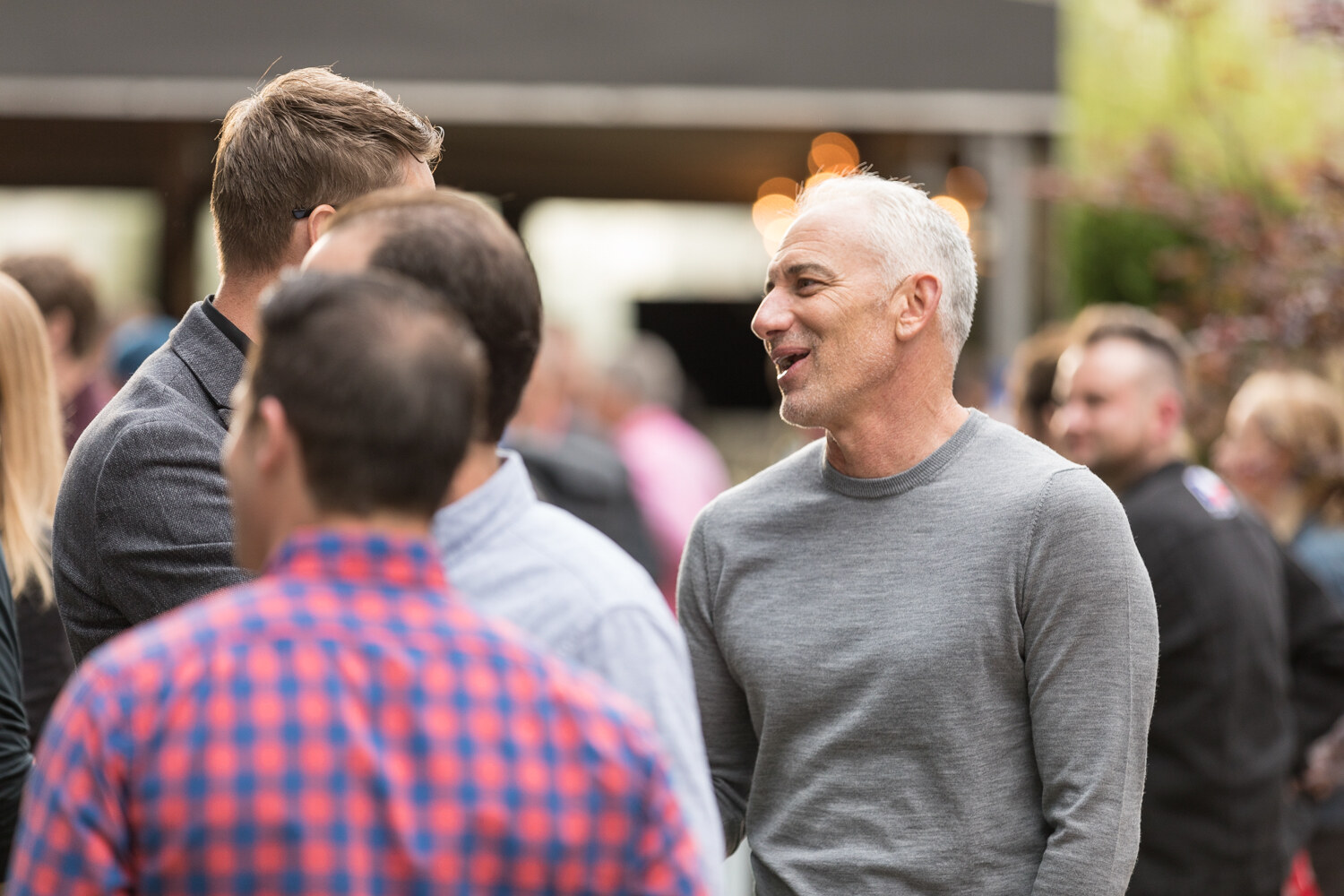 A silver-haired man in a gray sweater laughs and talks with a group of men outdoors.