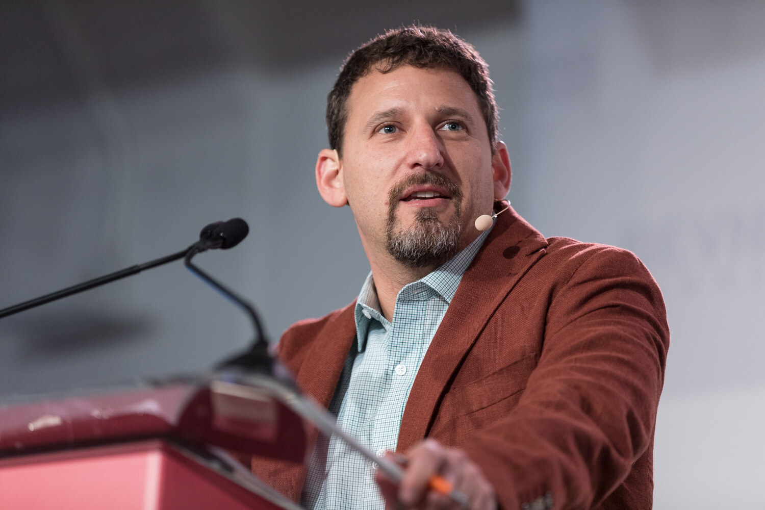 Close-up of a bearded man in a rust blazer and light checked shirt speaking beside a lectern microphone.