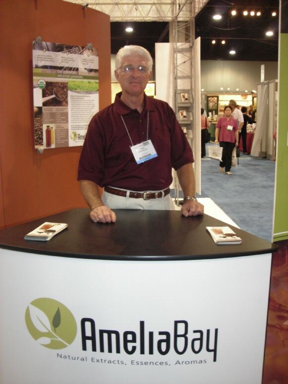 A gray-haired man with glasses and a maroon polo rests his hands on an AmeliaBay counter holding brochures, with a poster and the show floor behind him.