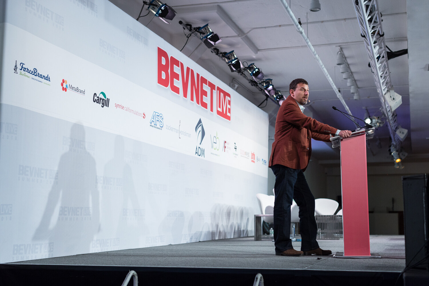 Wide view of a man in a rust blazer speaking at a red BevNET Live lectern on a long stage with sponsor logos on the backdrop.