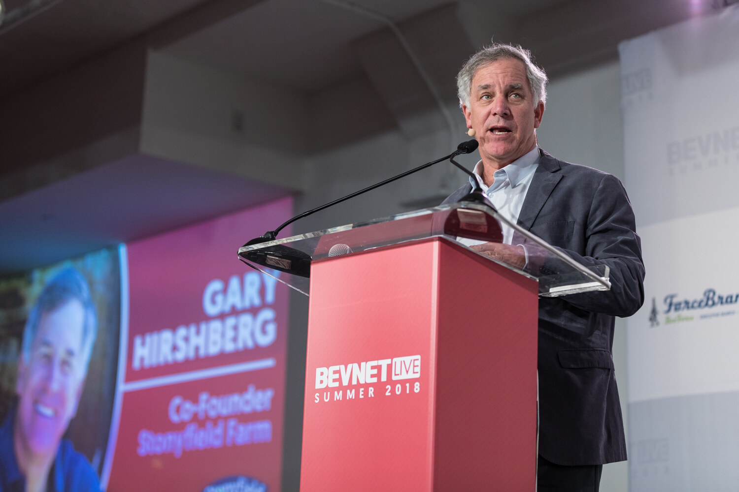 A man in a gray blazer speaks at a red lectern beside a large screen reading Gary Hirshberg, Co-Founder, Stonyfield Farm.