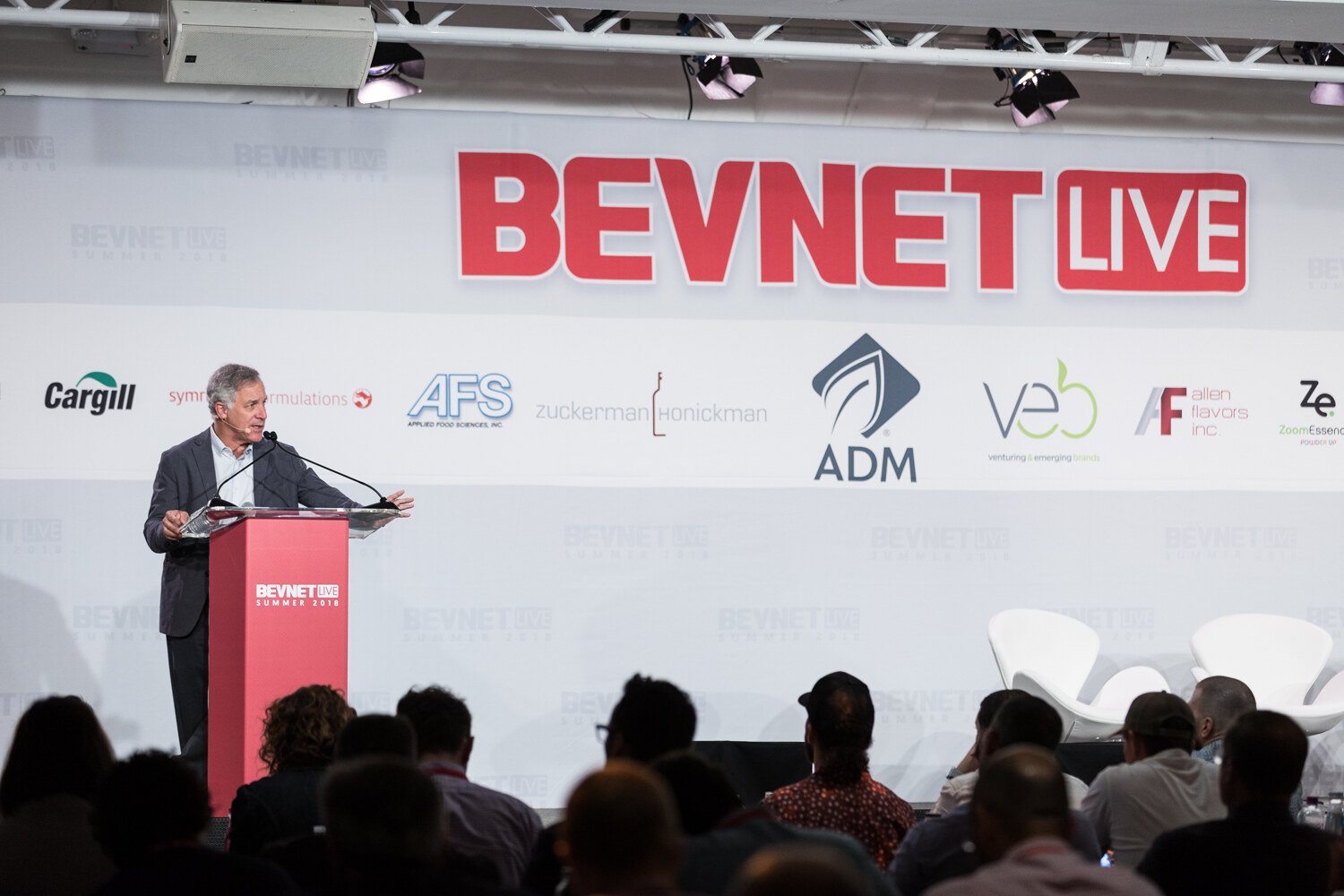 Wide view of a man at a red BevNET Live lectern in front of silhouetted audience members and the sponsor backdrop.