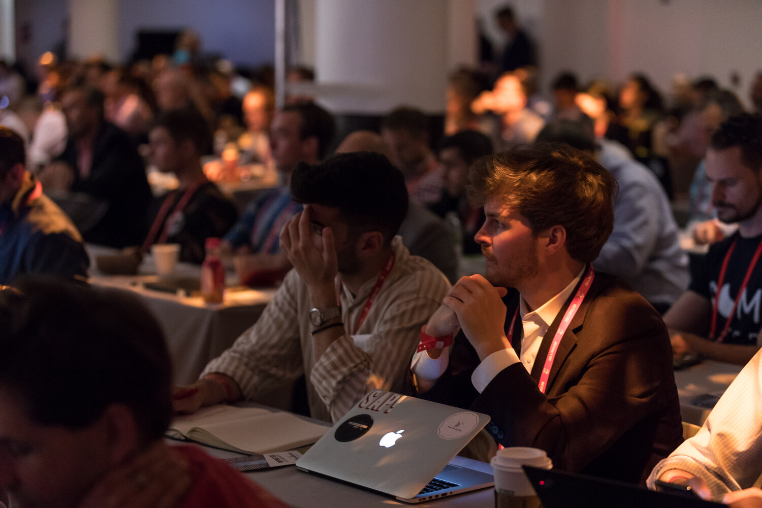 Seated attendees at tables with laptops and notebooks looking toward the stage, lit by warm room light.