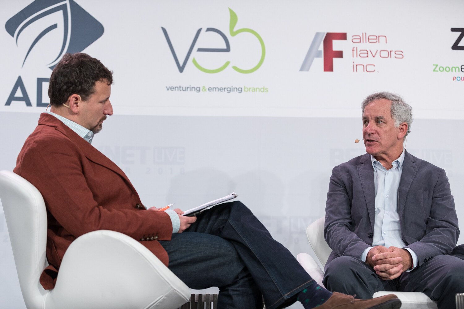 Two men in conversation on stage; one in a rust blazer holds notes while the other sits with hands clasped.