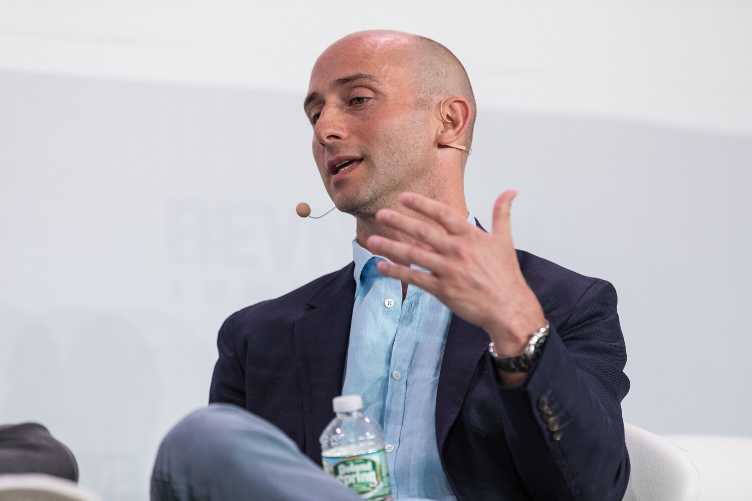 Close-up of a bald man in a navy blazer and light blue shirt gesturing with one hand, with a water bottle in the foreground.