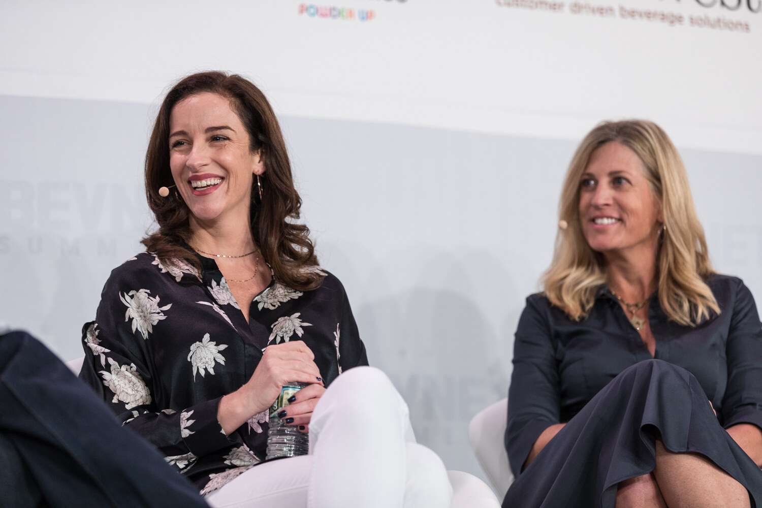 Two women in dark outfits seated on stage, both smiling and looking toward a speaker out of frame.