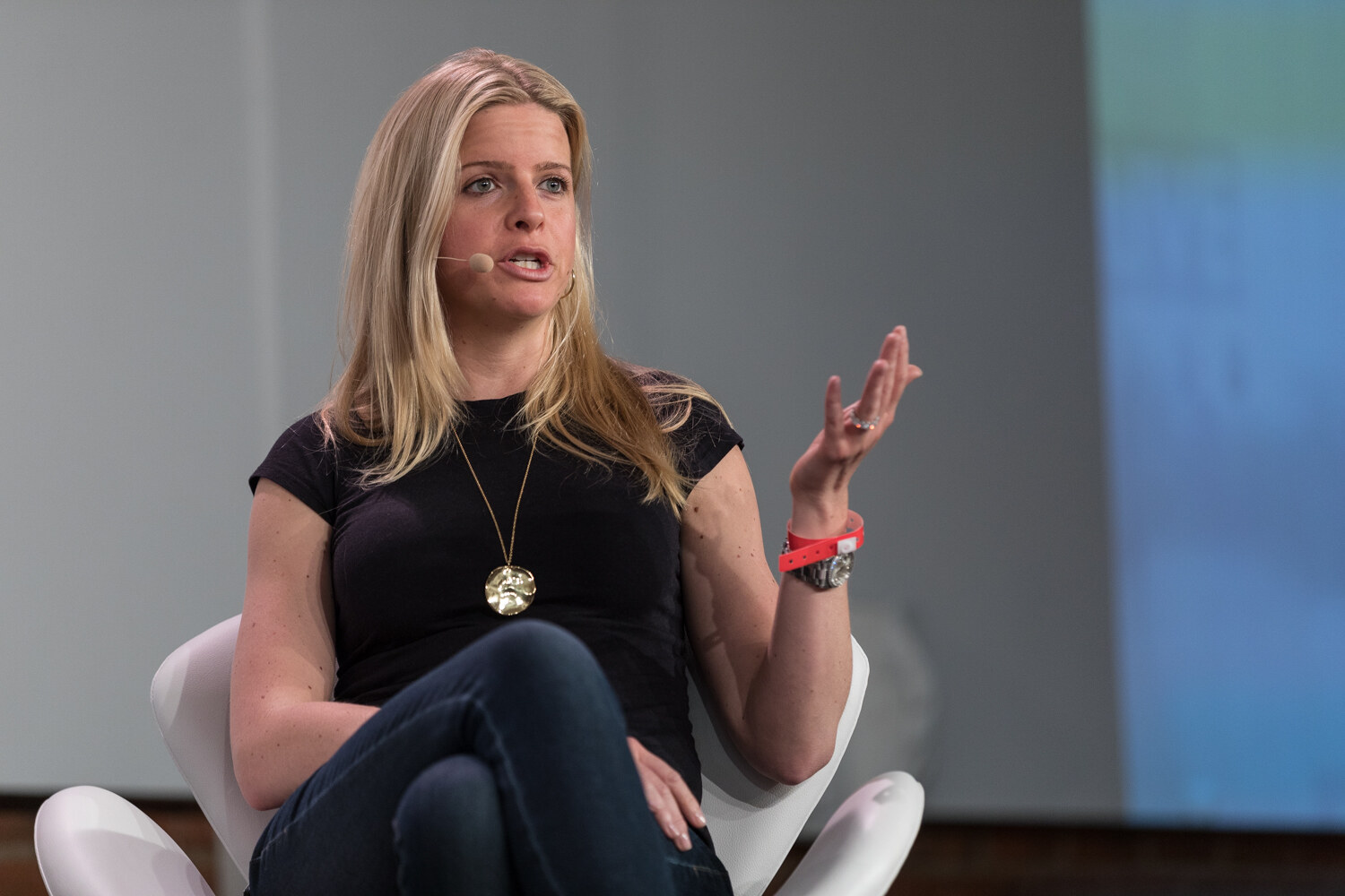 A blonde woman in a black top and jeans sits in a white chair and gestures with one hand.