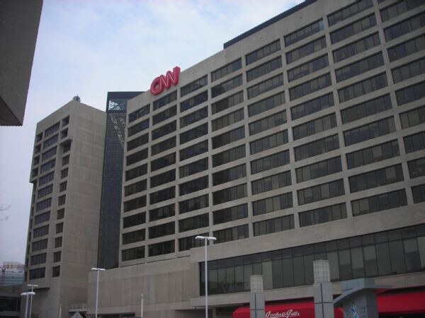 Exterior of a multi-story office building with a red CNN sign on its upper facade, under a pale sky, with street lamps in the foreground.