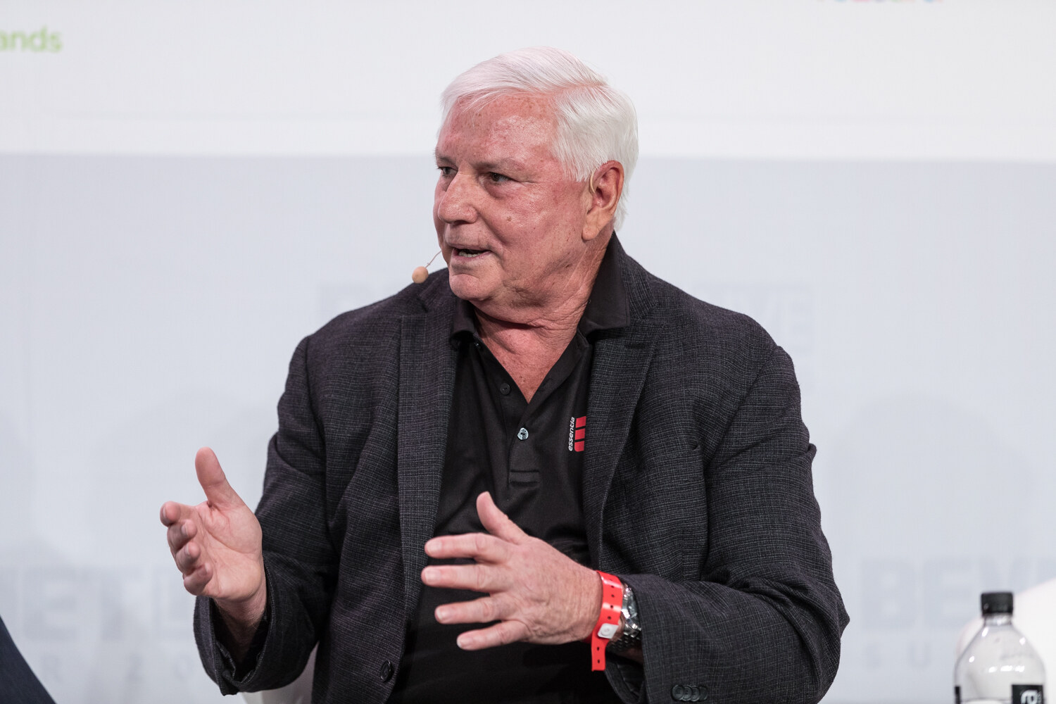 A white-haired man in a dark jacket and black polo gestures with both hands while seated on stage.