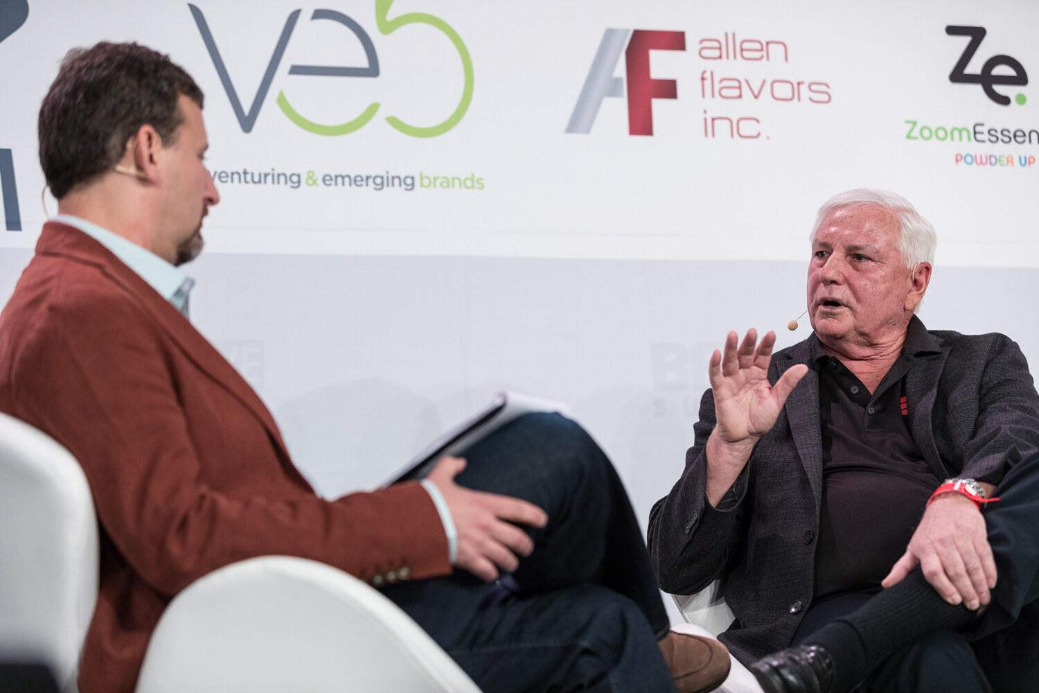 A man in a rust blazer holds a tablet while a white-haired man across from him raises a hand to make a point.