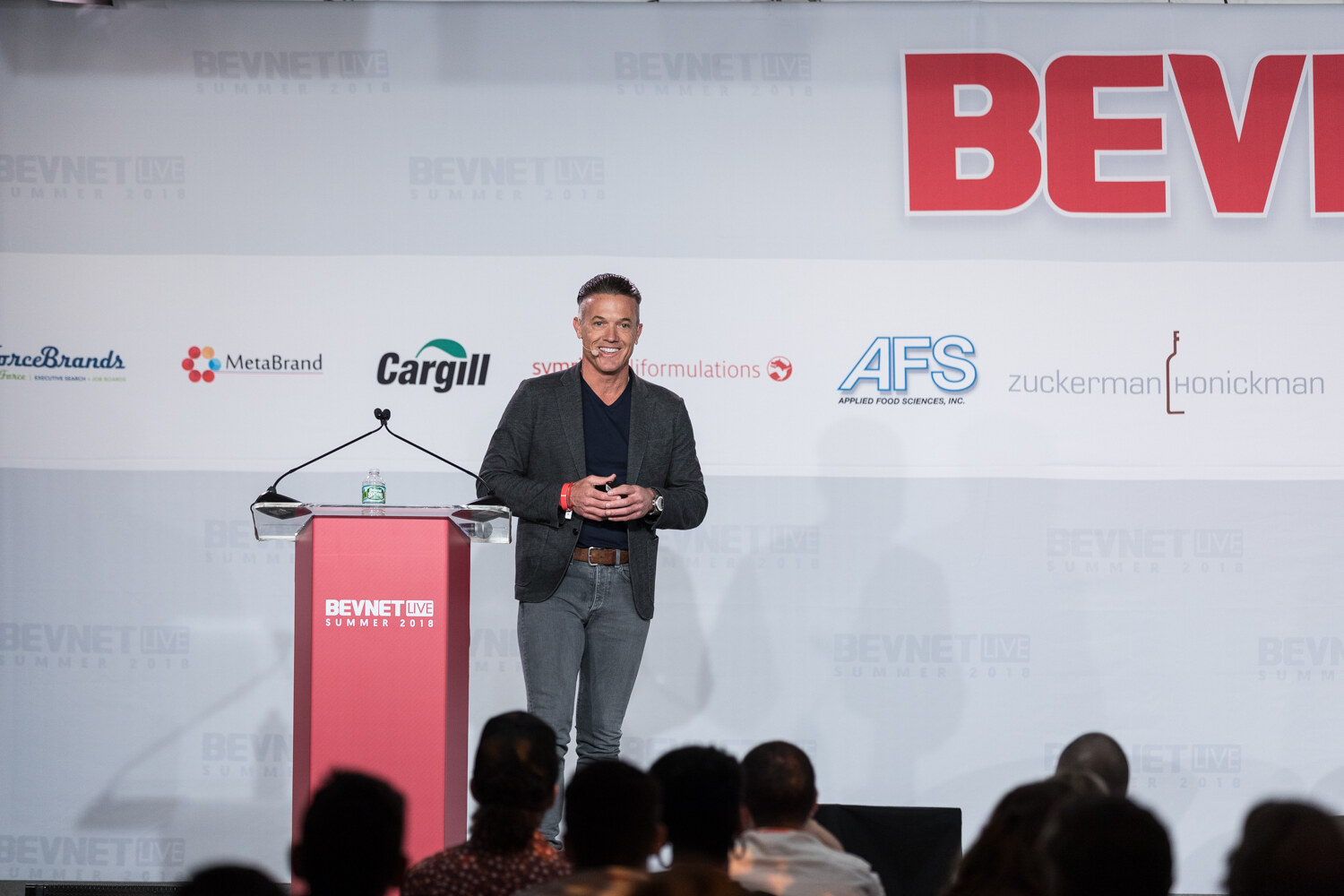 A man in a dark jacket stands at a red BevNET Live lectern in front of the sponsor backdrop with audience heads in the foreground.