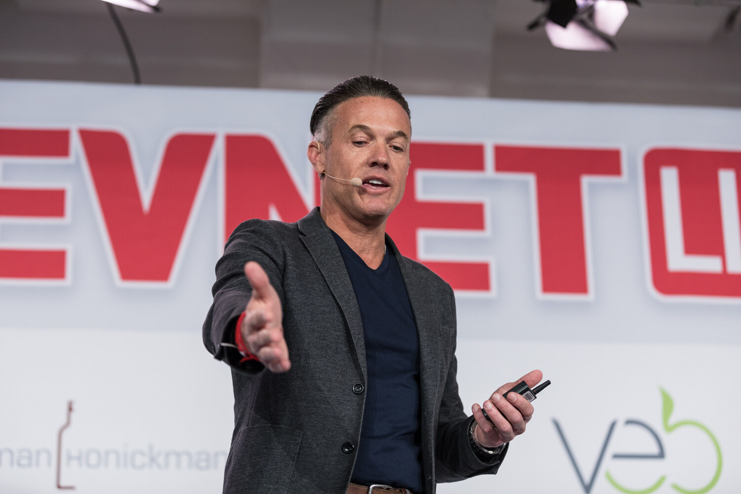 A man in a gray blazer and navy shirt gestures with an open hand in front of the large BevNET Live sign.