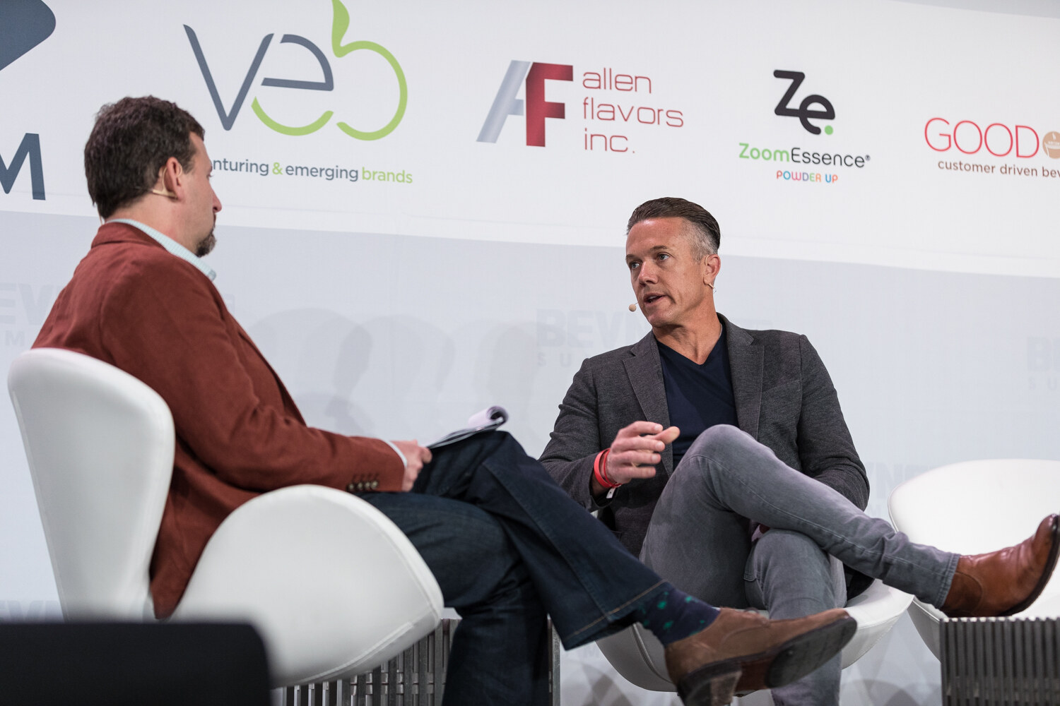 Two men seated in white swan chairs on stage in conversation, with Veb, Allen Flavors and ZoomEssence logos behind them.