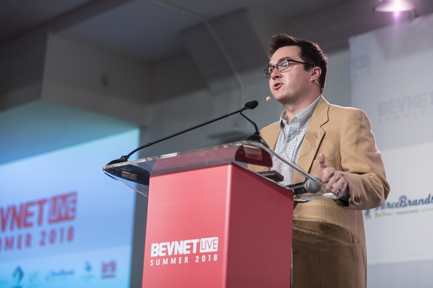 A man in glasses and a tan jacket speaks at a red BevNET Live Summer 2018 lectern.