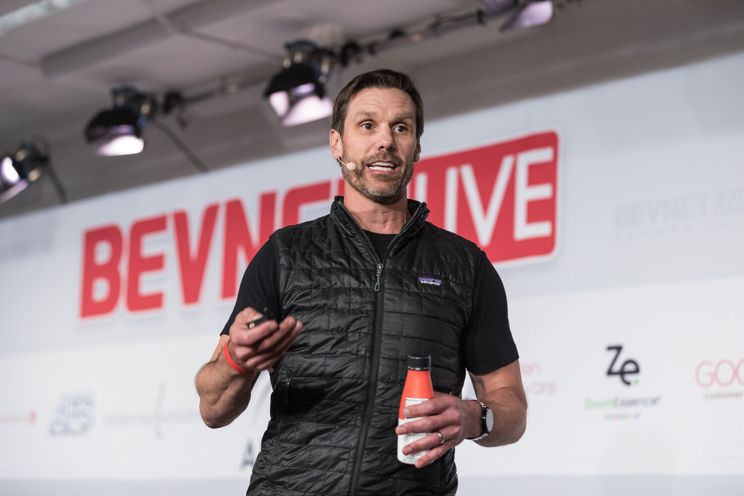 A man in a black puffer vest holds an orange and white bottle and a clicker in front of the BevNET Live backdrop.