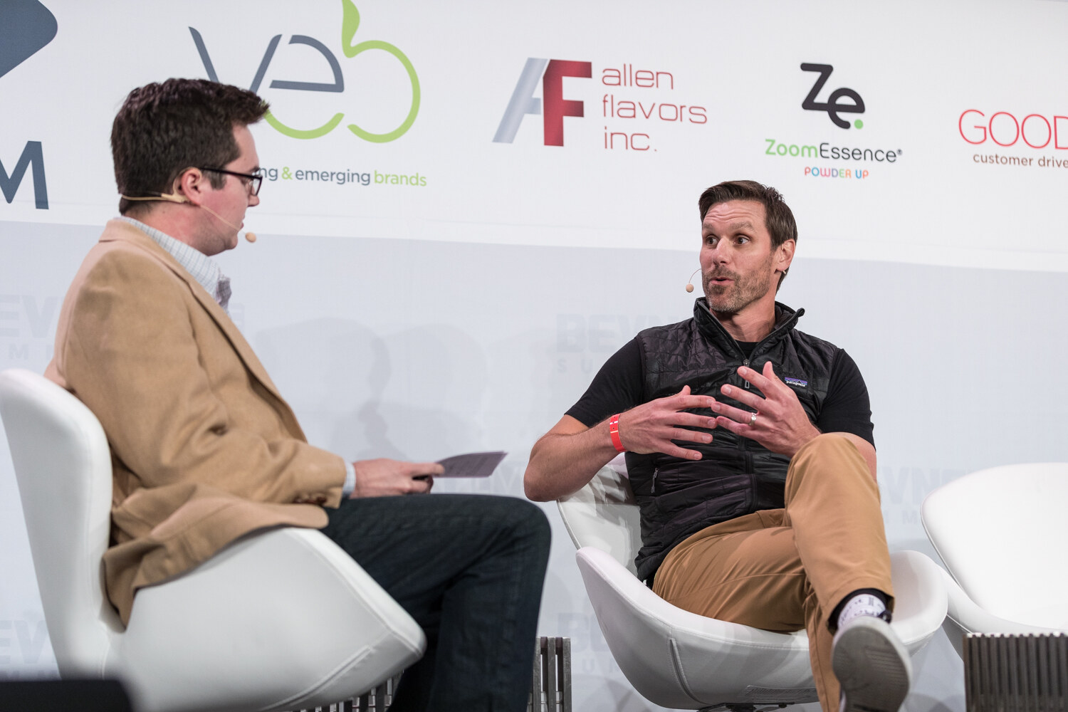 Two men seated in white chairs on stage in conversation; one wears a tan jacket, the other a black vest and khakis.