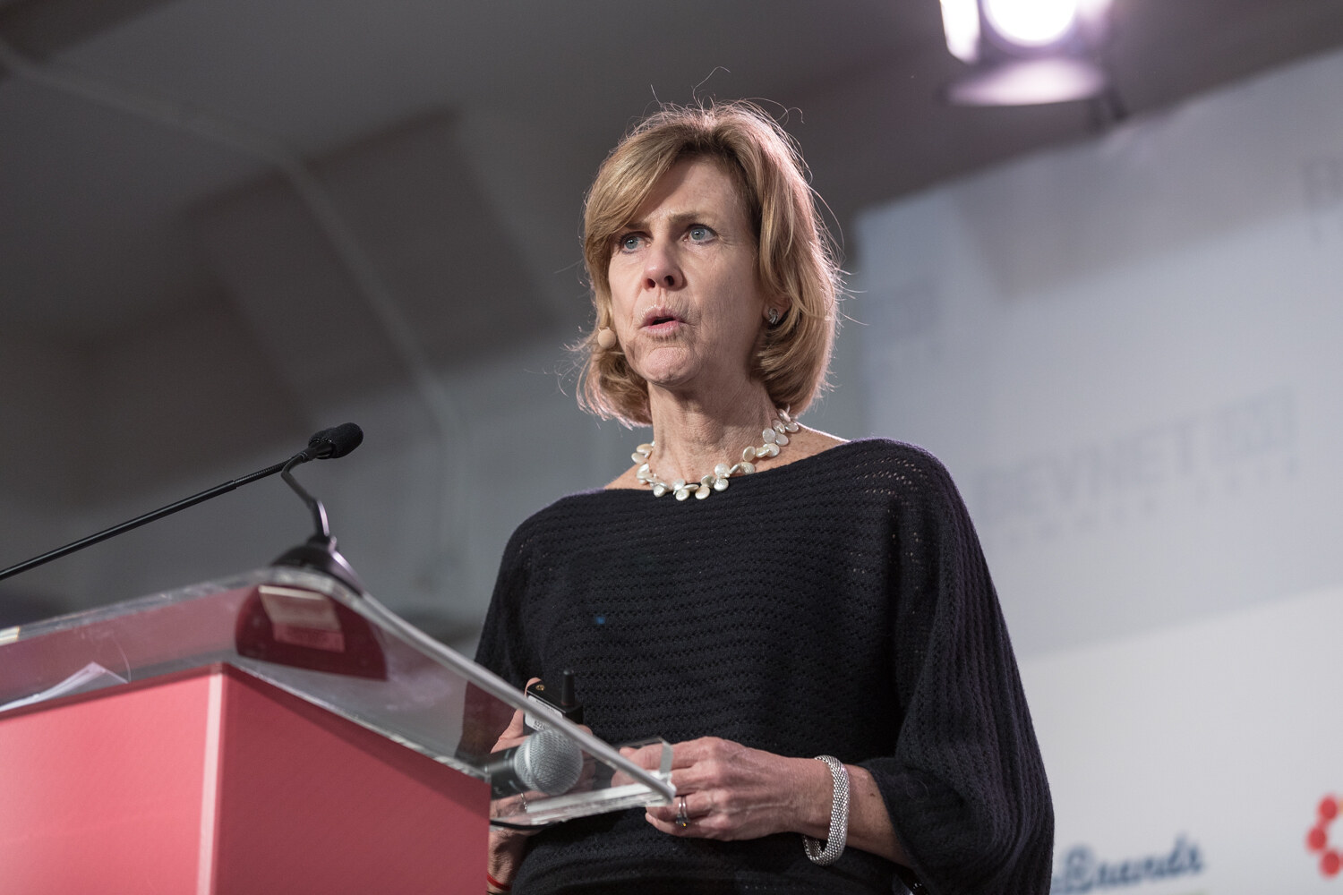 A woman in a dark knit top speaks at a red lectern with a microphone and a slide clicker in her hand.