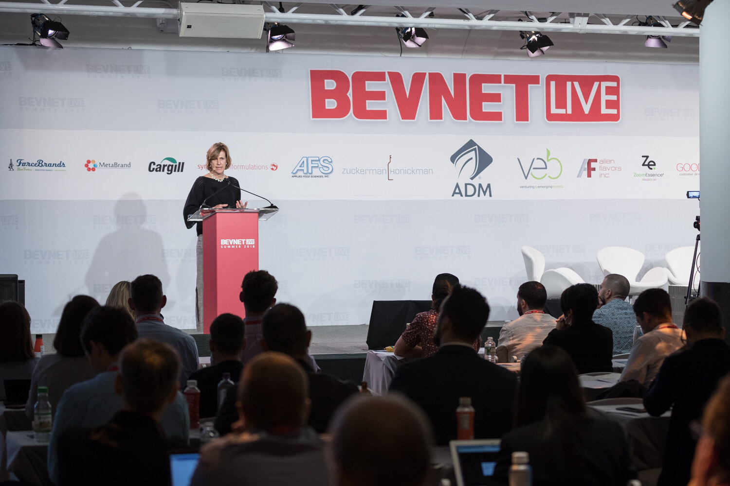 Wide view of a room with seated attendees facing a stage, a woman at a red lectern and the BevNET Live sponsor backdrop.