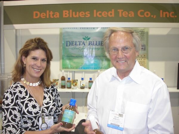 A smiling woman and a man with glasses hold bottles of tea at the Delta Blues Iced Tea Co., Inc. booth, with bottles on a shelf behind them.