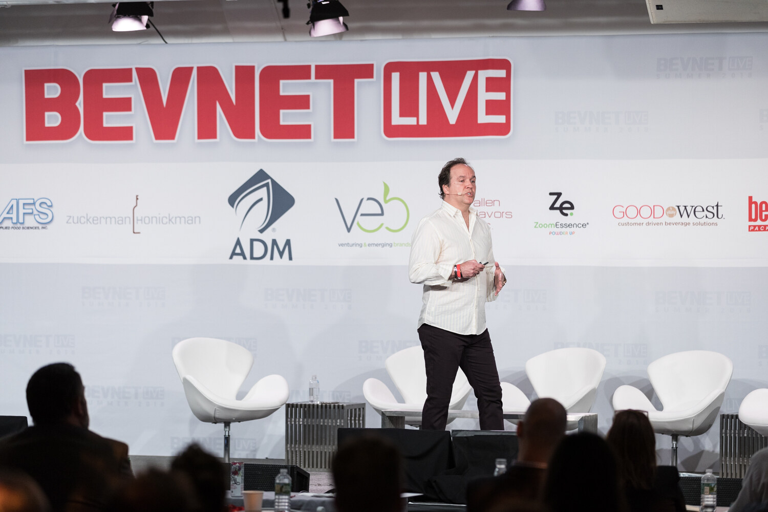 Wide view of a man in a white shirt and dark pants walking across a stage with white swan chairs and the BevNET Live backdrop.