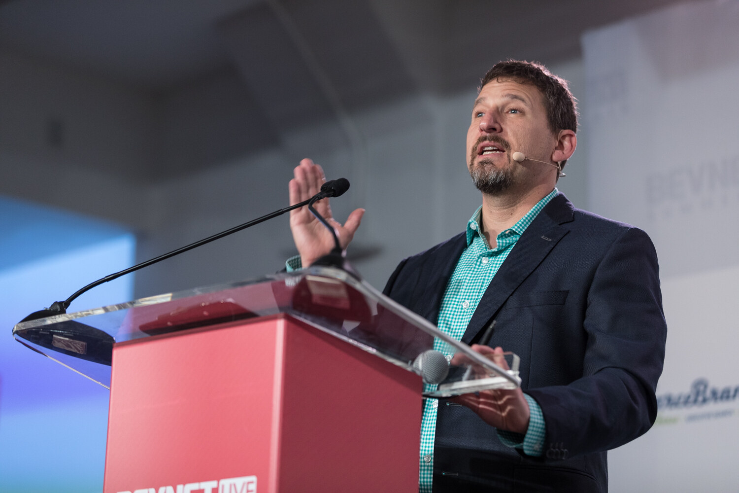 A man in a teal shirt and navy blazer gestures at a red BevNET Live lectern.