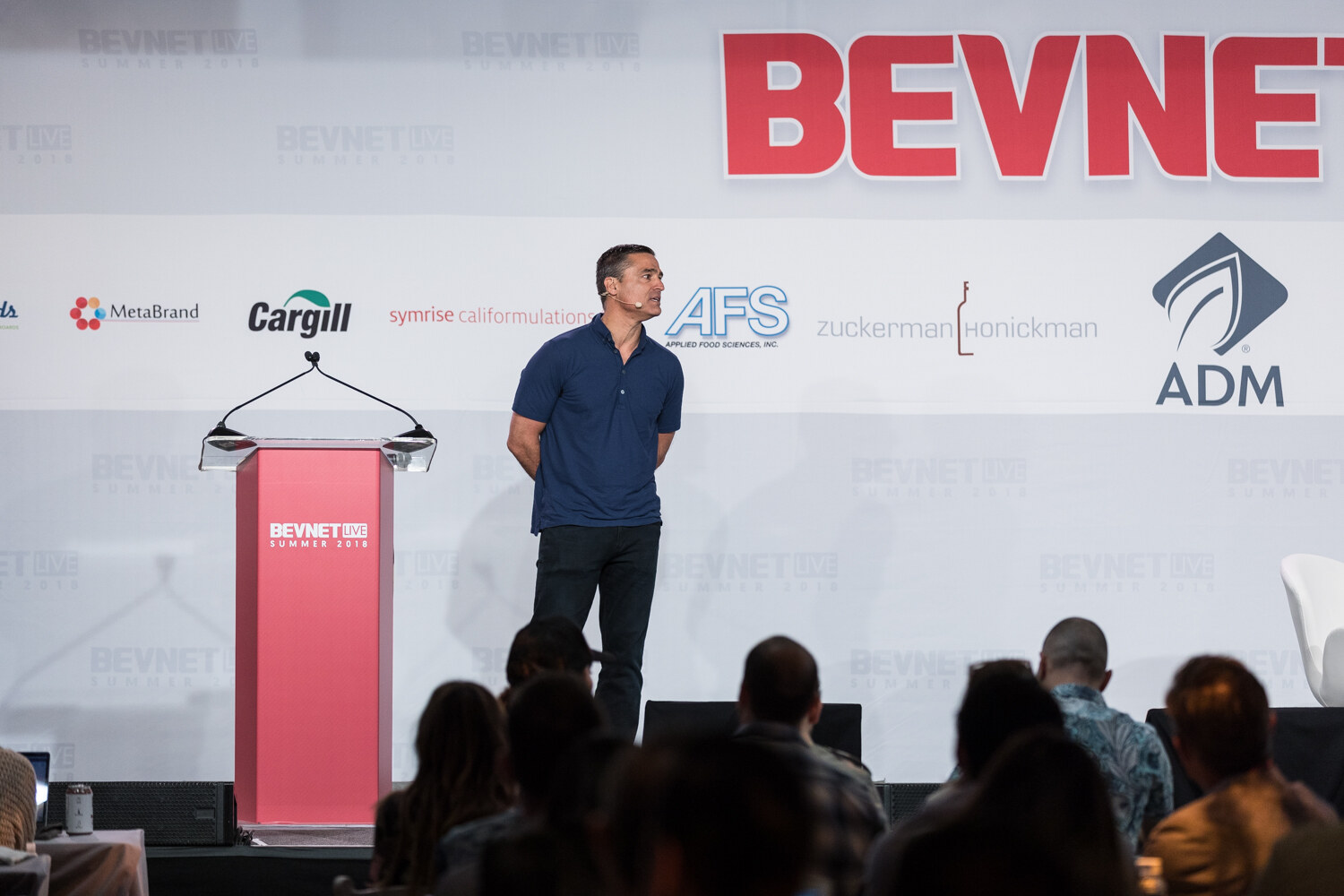 A man in a navy polo stands with his hands behind his back beside an empty red lectern with the BevNET Live backdrop behind him.