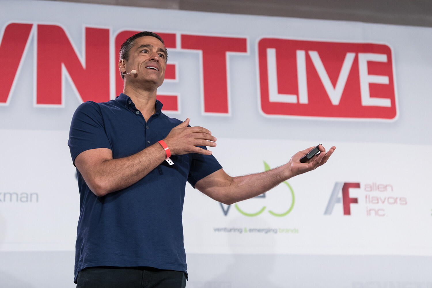 A man in a navy polo looks up and gestures with an open hand in front of the red BevNET Live sign.