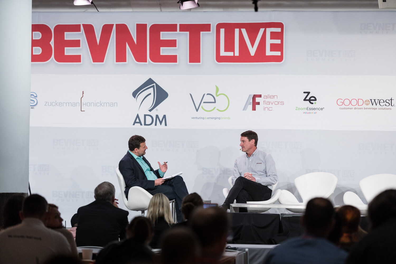 Wide view of two men seated on stage in white chairs in front of the BevNET Live backdrop with audience silhouettes in the foreground.