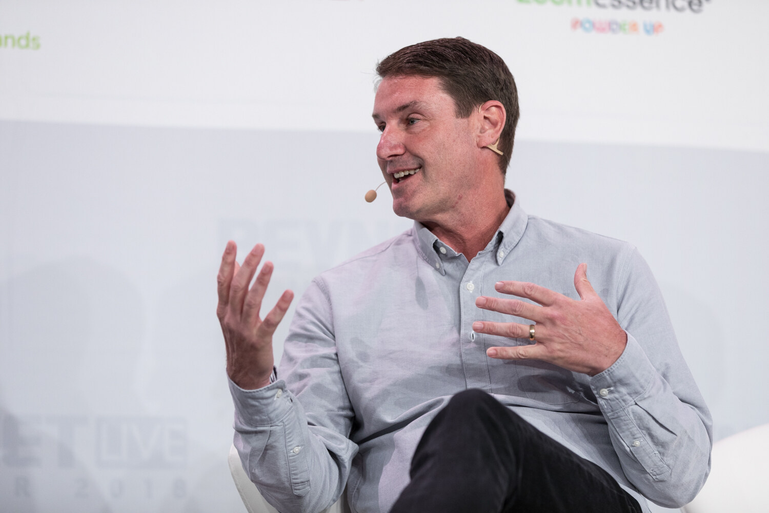 A man in a light gray shirt with a small Chobani logo gestures with both hands while seated and speaking.