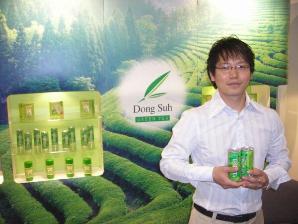 A man with glasses holds two green cans in front of a Dong Suh Green Tea backdrop of tea fields, beside a lit shelf of green tea products.