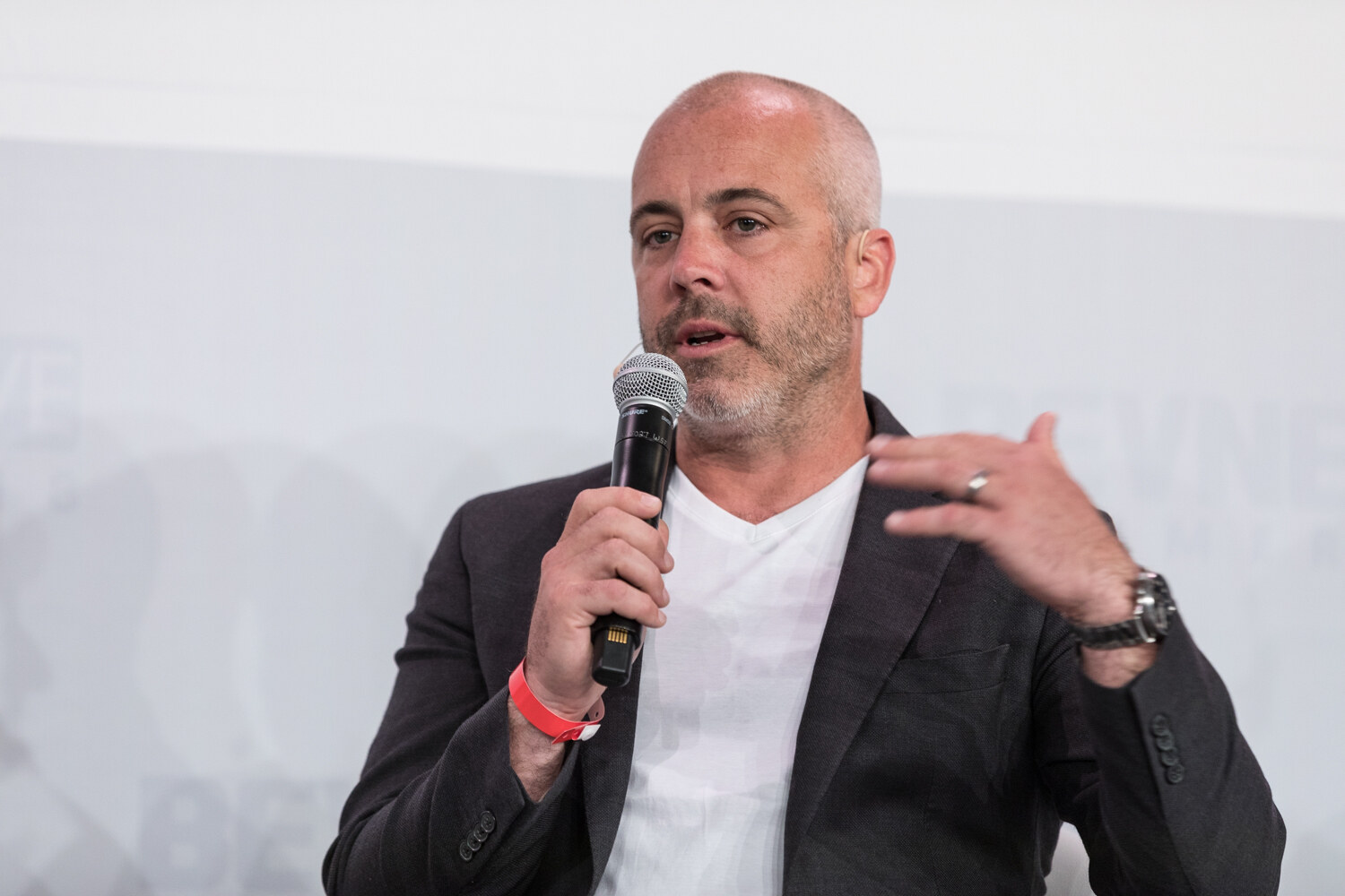 A bald man in a dark blazer and white shirt speaks into a handheld microphone and gestures with his free hand.