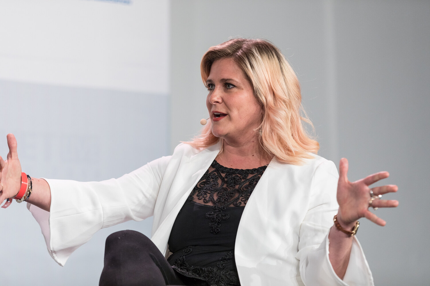 A woman with blonde hair in a white blazer and black lace top extends both arms while speaking.