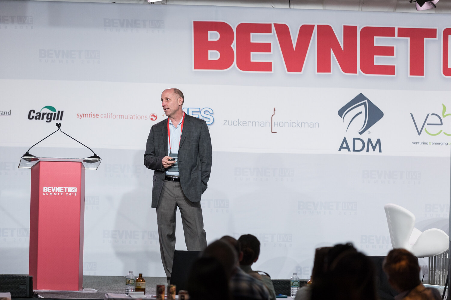 A man in a dark blazer and gray pants wearing a lanyard stands on stage beside a red BevNET Live lectern.