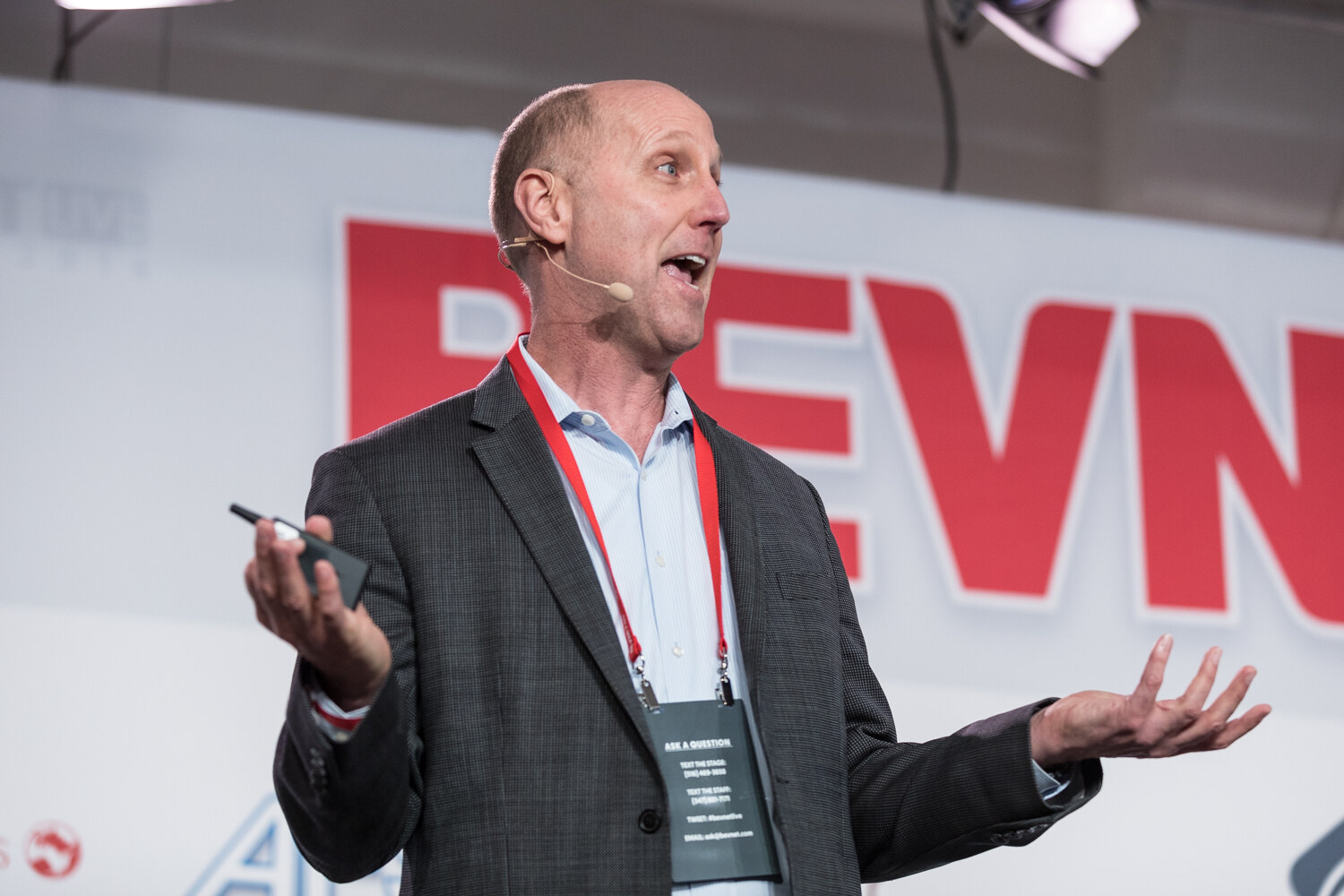 A man in a gray checked blazer and lanyard speaks with both hands open in front of the large BevNET Live sign.