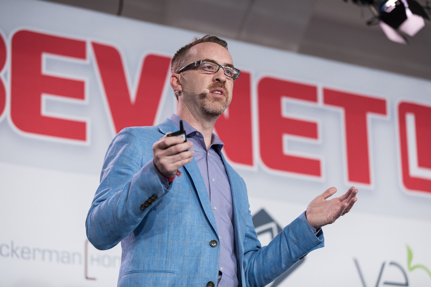Close view of a bearded man in glasses and a light blue blazer gesturing with one open hand in front of the red BEVNET LIVE letters.