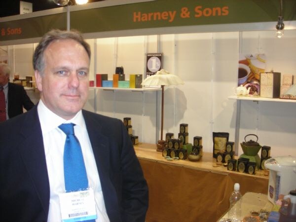 A man in a dark suit and blue tie wearing a show badge stands in front of the Harney & Sons booth, where tins of tea, a teapot and boxes sit on shelves.