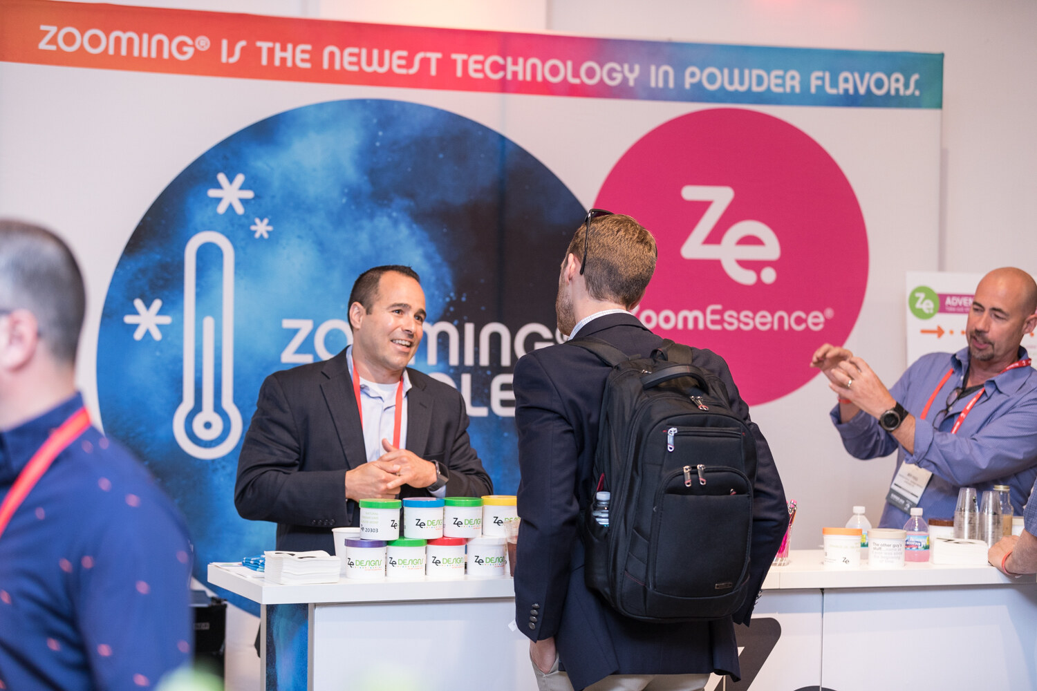 A man in a dark blazer behind a white table with tubs of powder in front of a ZoomEssence banner, with an attendee in a backpack in the foreground.