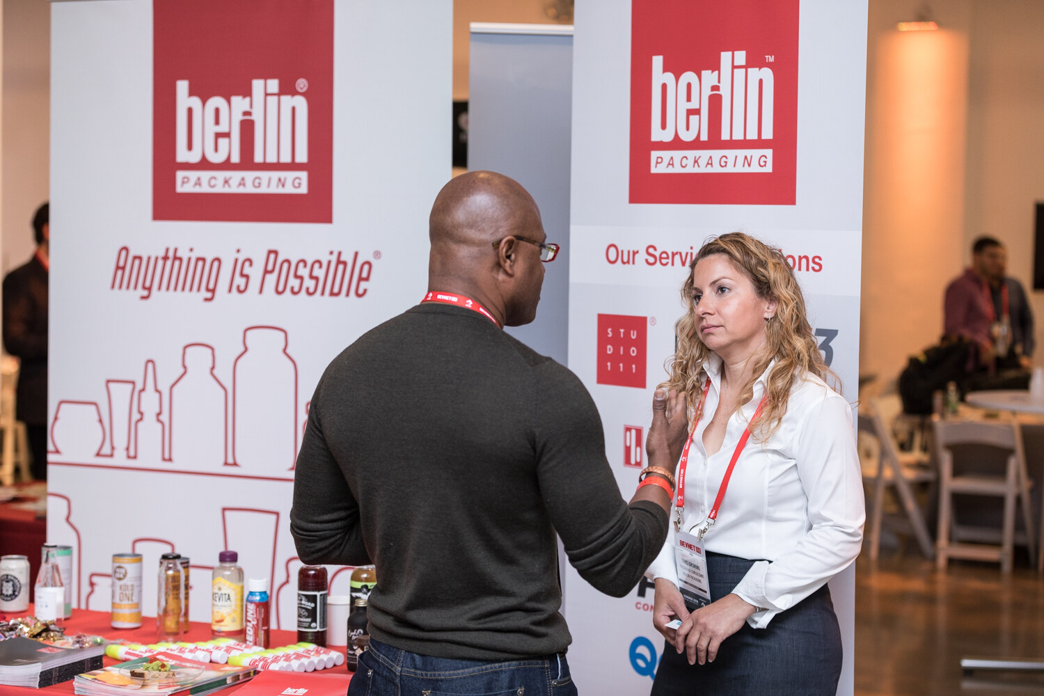A man and a woman talk in front of Berlin Packaging banners reading Anything is Possible, with sample bottles on a red table.