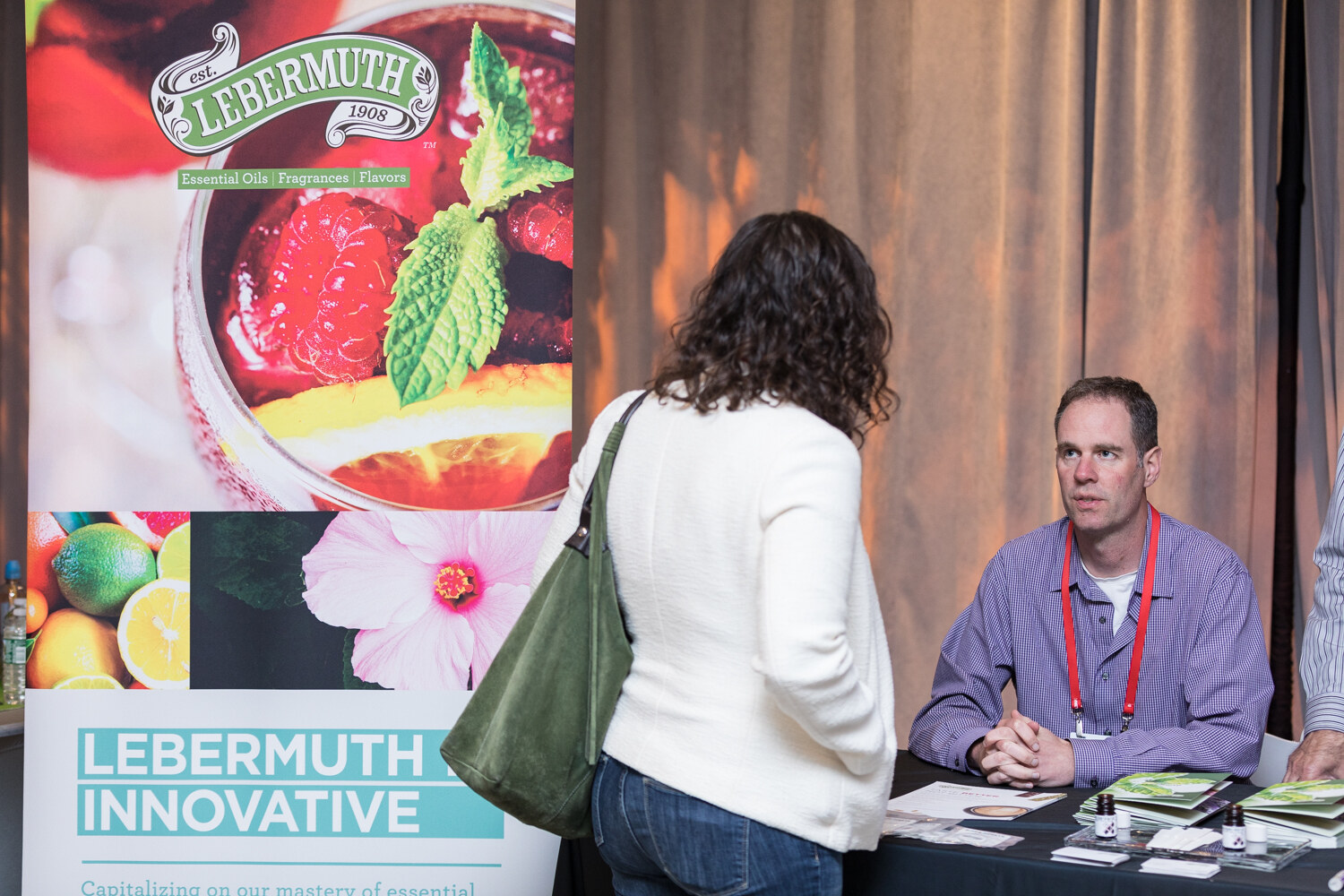 A man wearing a lanyard sits at a table in front of a Lebermuth Innovative banner while a woman with a green bag talks with him.