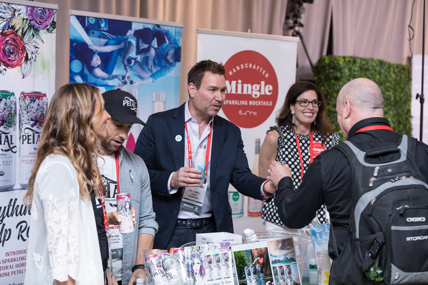 A group of five people talk at a booth with a Mingle Handcrafted Sparkling Mocktails banner and cans on the table.