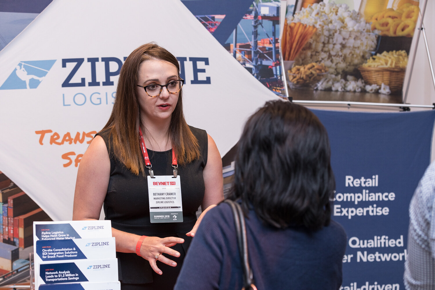 A woman wearing a Zipline Logistics badge speaks with an attendee in front of a Zipline banner about retail compliance expertise.