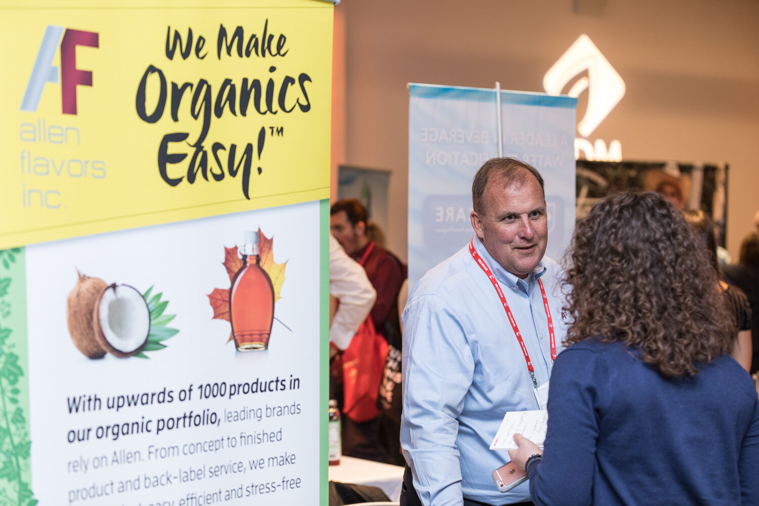 A man in a light blue shirt and lanyard talks with a woman beside an Allen Flavors banner reading We Make Organics Easy.