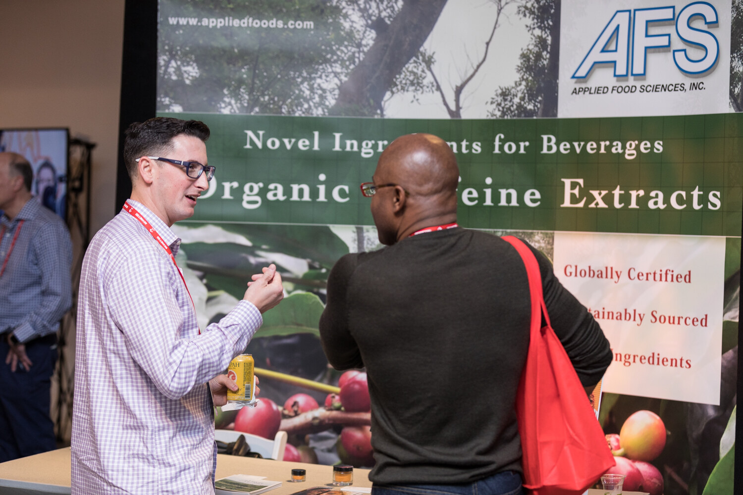 Two men talk in front of an Applied Food Sciences banner reading Novel Ingredients for Beverages, Organic Caffeine Extracts.
