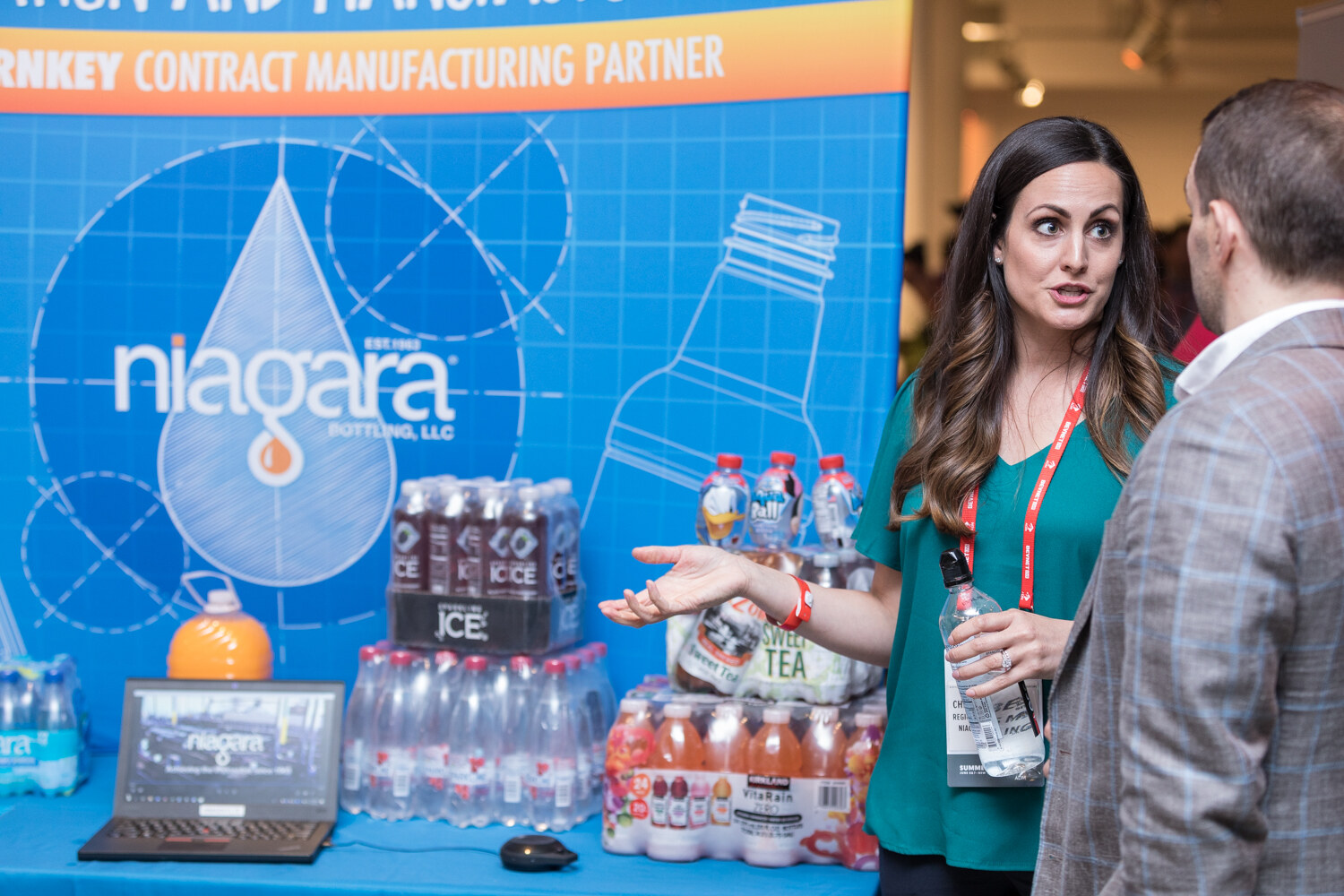 A woman in a teal top speaks with an attendee in front of a Niagara Bottling banner with bottled water and tea on display.