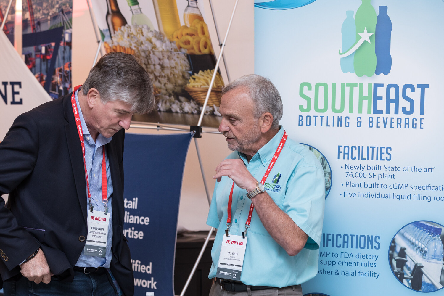 Two men wearing lanyards talk in front of a Southeast Bottling & Beverage banner.