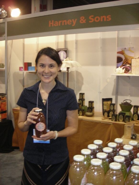 A smiling woman in a dark blouse holds a bottled drink at the Harney & Sons booth, with tea tins on a table behind her and rows of bottled tea in front.