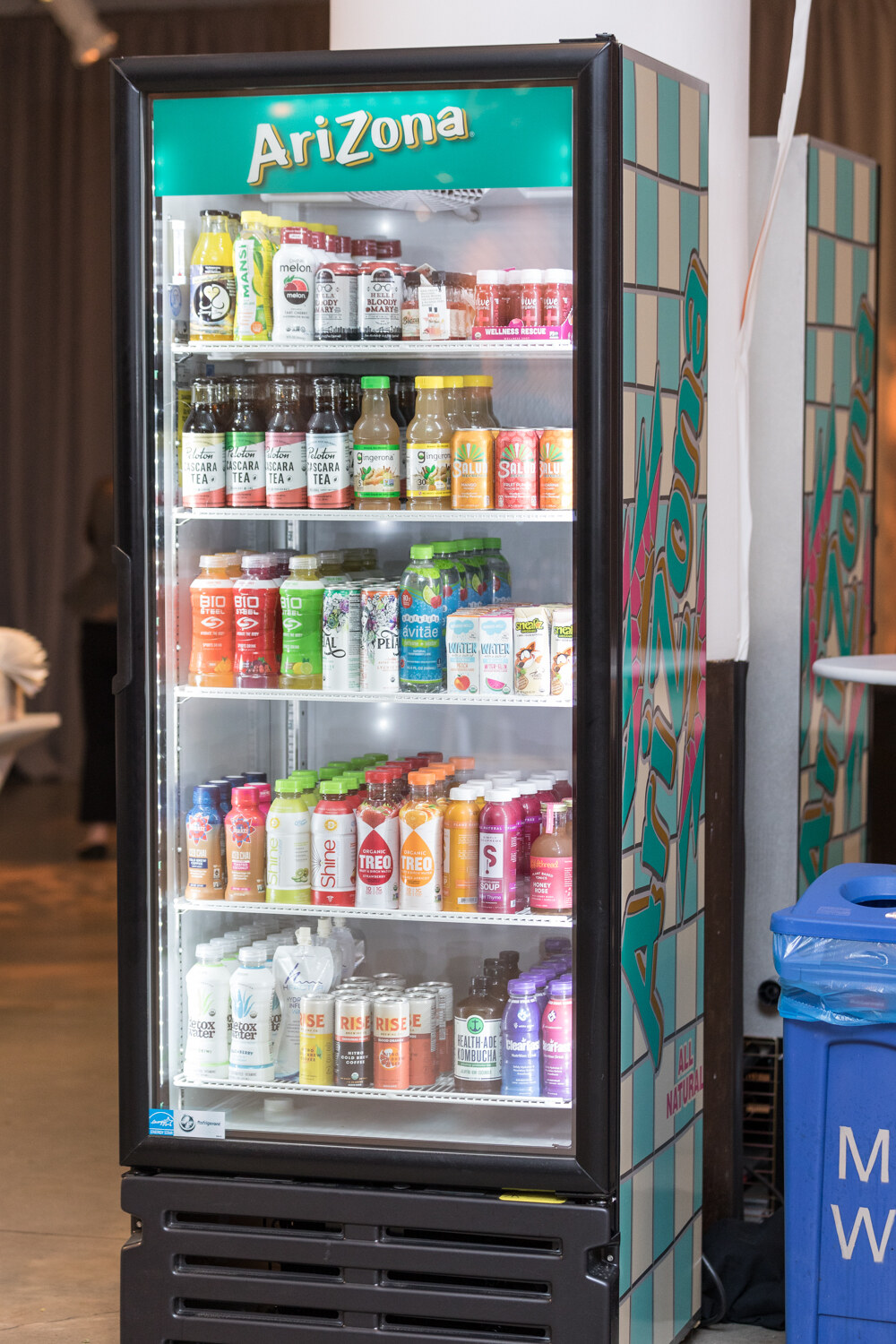 An upright glass-door cooler with an AriZona sign on top, filled with bottled and canned drinks.