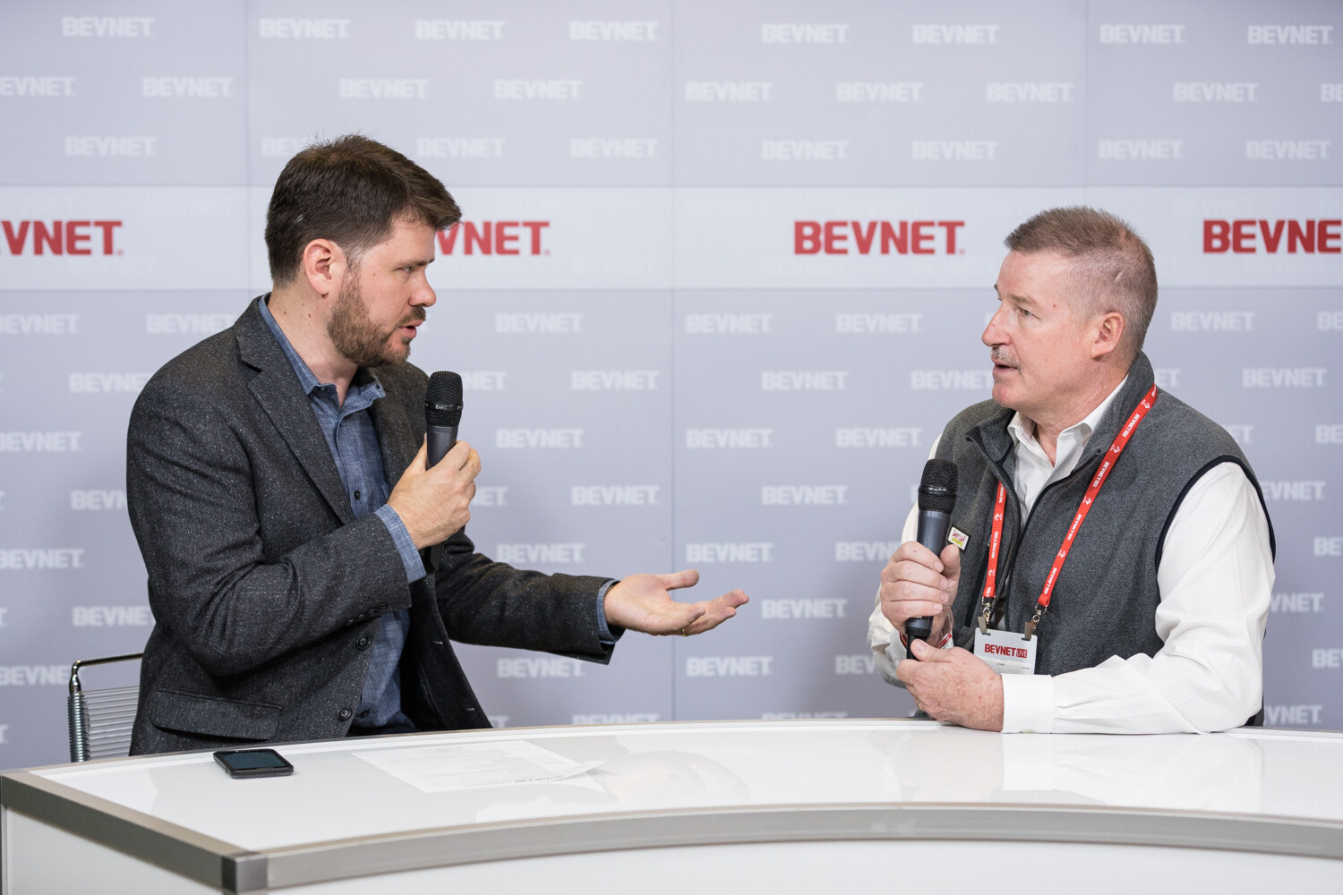 A bearded host and a man in a gray vest with a lanyard talk with microphones at a white desk.