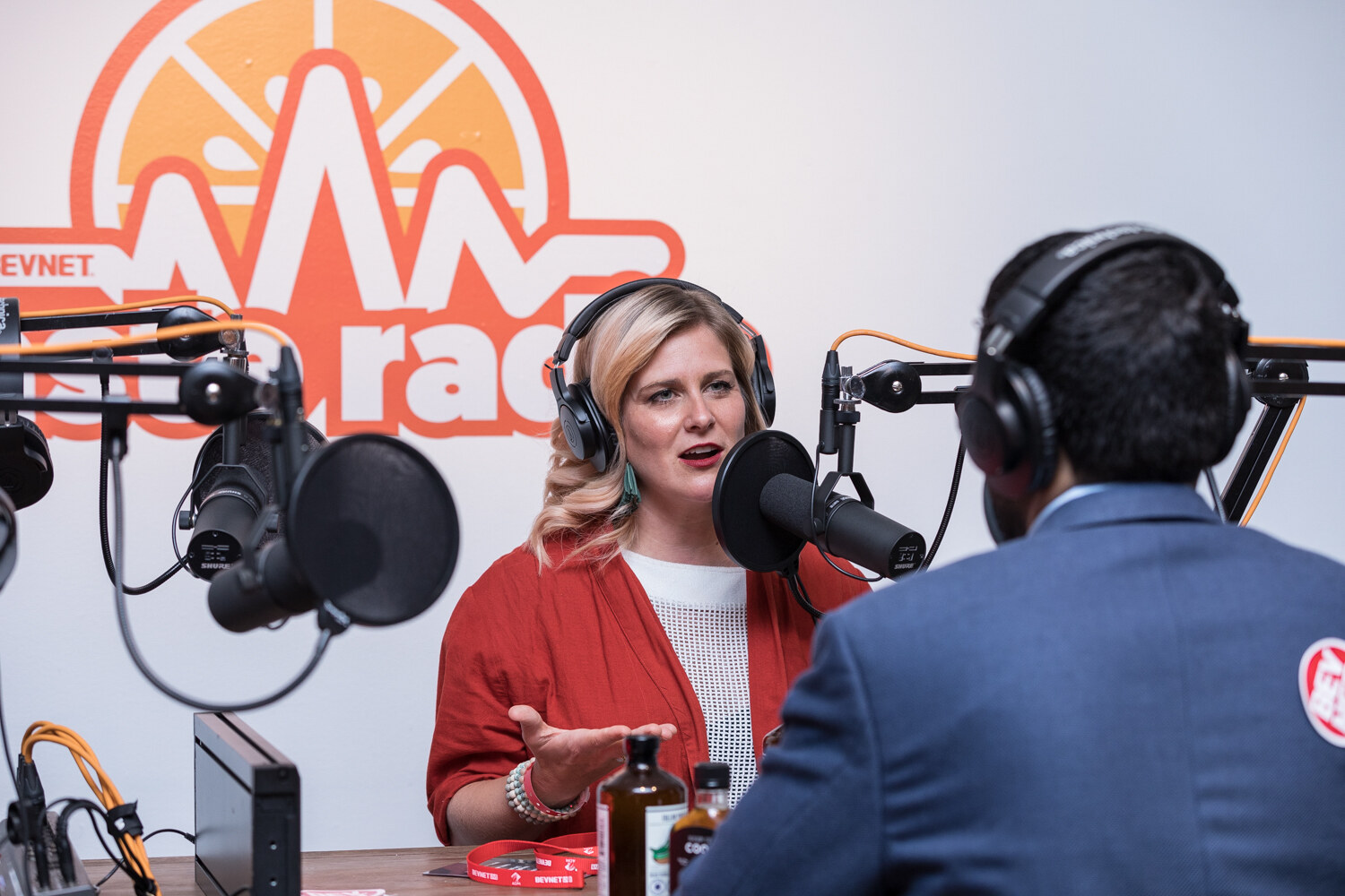 A woman in a red cardigan and a man seen from behind wear headphones at microphones in front of an orange Taste Radio logo.