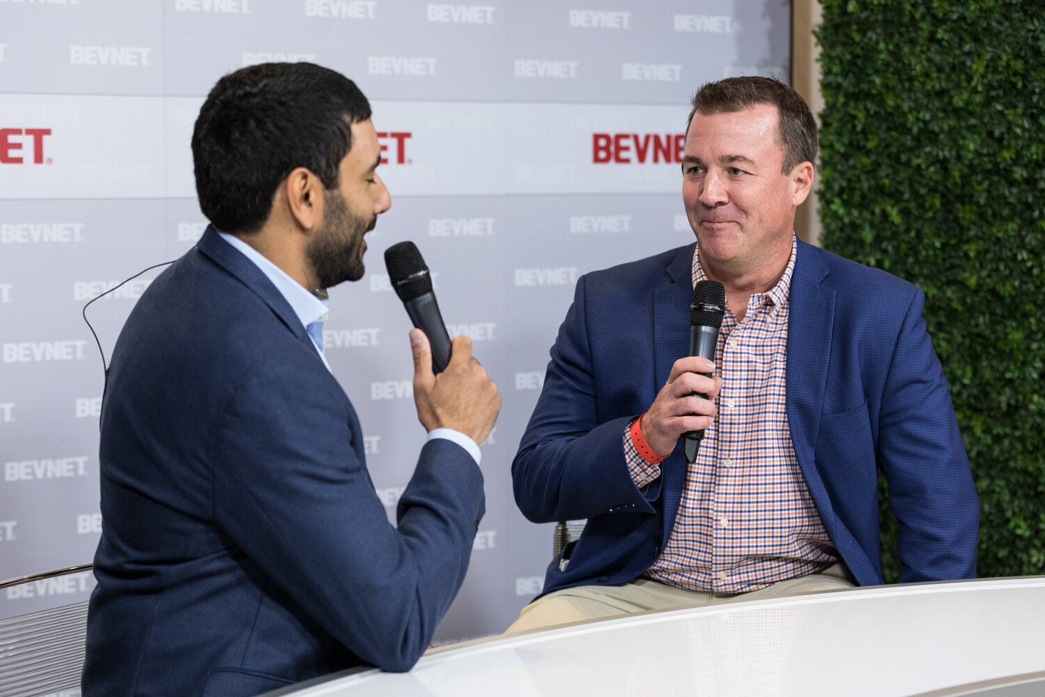 Two men in suit jackets speak into handheld microphones at a white desk in front of a gray BevNET-patterned backdrop.